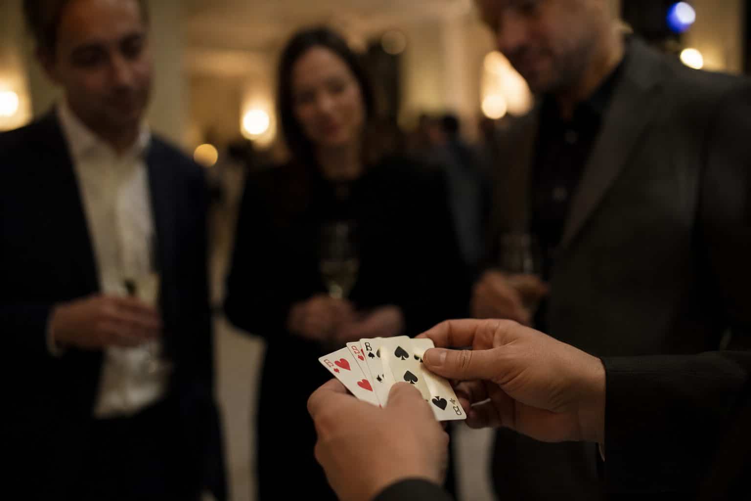 Hände mit aufgefächerten Spielkarten auf Brusthöhe, drei Gäste eines Stehempfangs schauen darauf