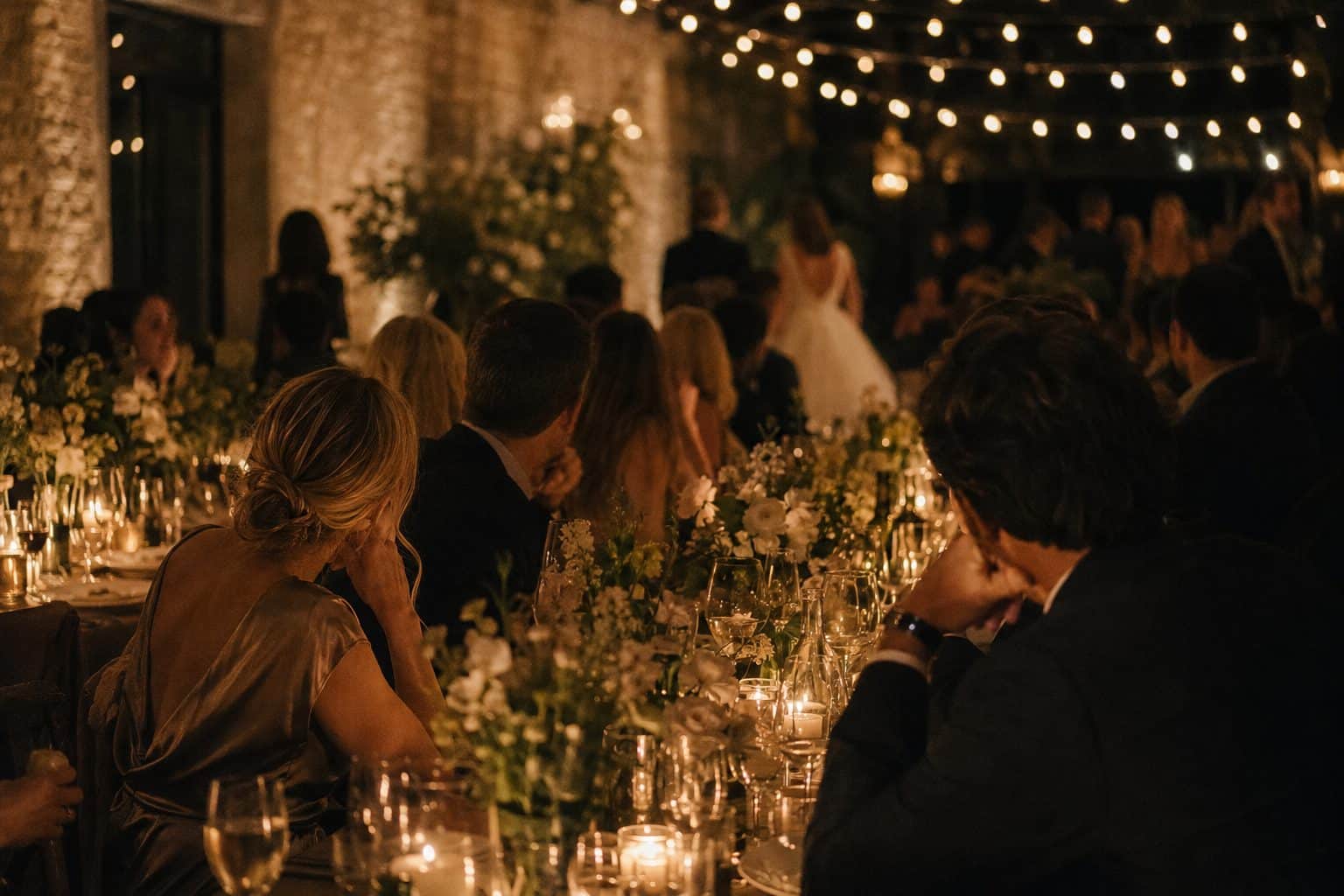 Lange Hochzeitstafel am Abend mit niedrigen Kerzen und lose gesteckten Blumen, Gäste im Gespräch von hinten gesehen